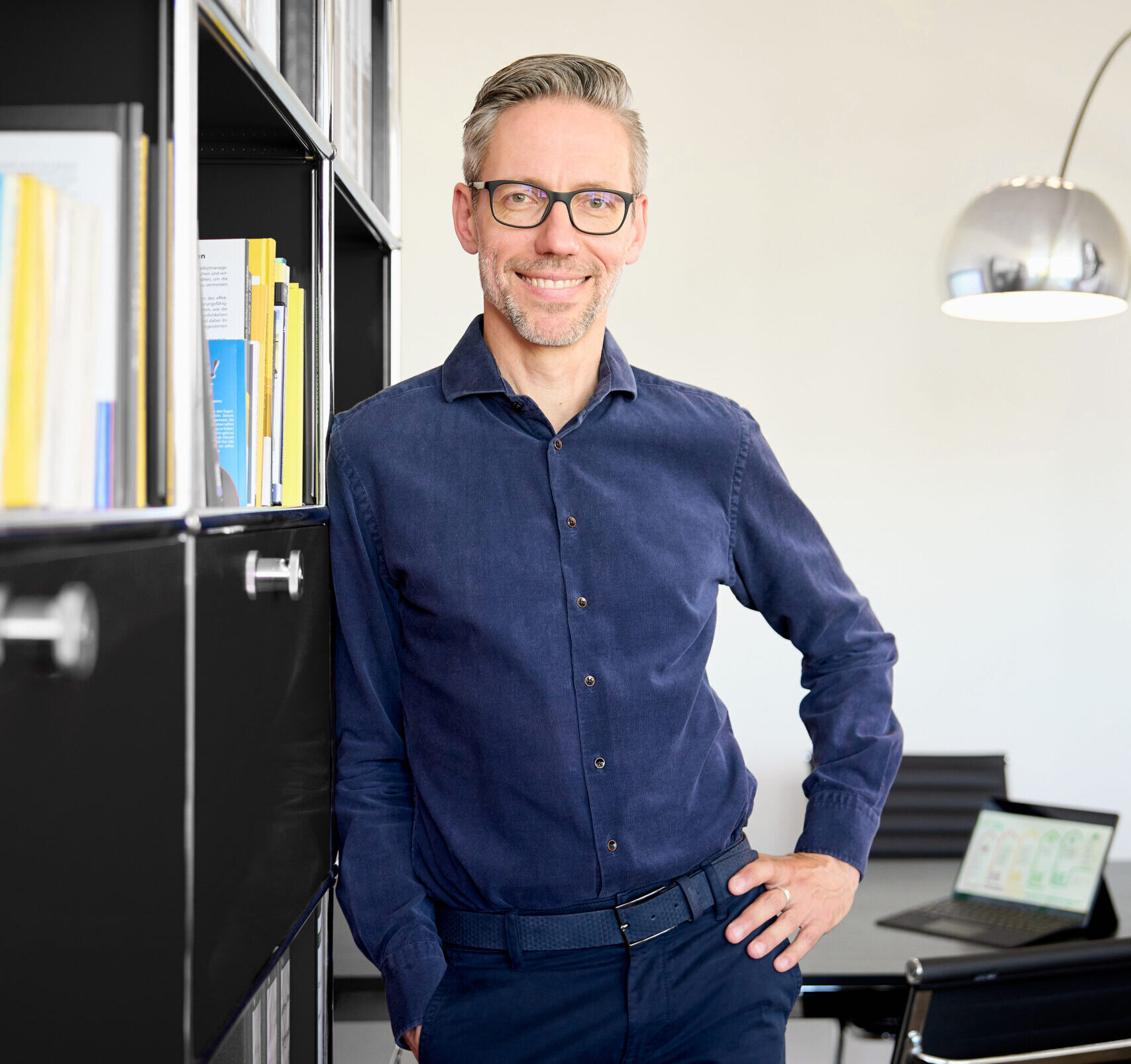 Energieberater München lehnt lächelnd an einem Regal in einem modernen Büro mit einem Laptop im Hintergrund.