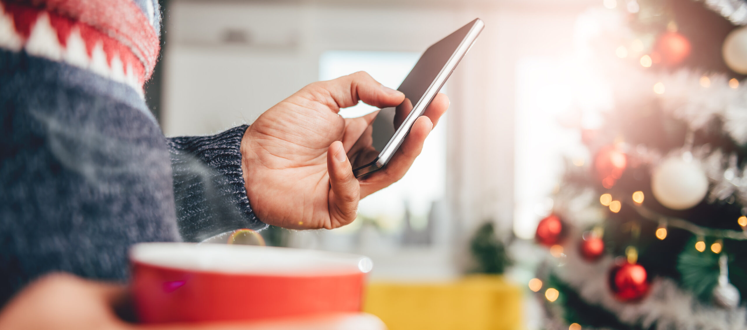 Hand mit Smartphone und Kaffeetasse vor einem Weihnachtsbaum – Energieberatung München auch zur Weihnachtszeit