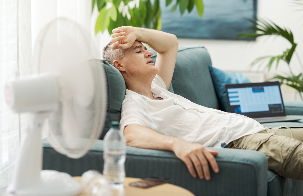 Mann liegt erschöpft auf dem Sofa vor einem Ventilator – Hitze im Sommer im Zuhause vermeiden